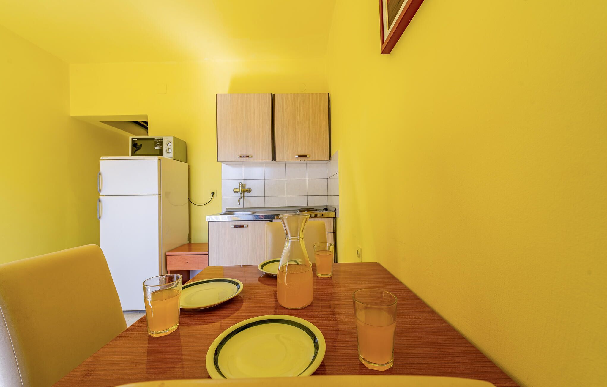 Une cuisine compacte avec un réfrigérateur blanc, des placards en bois et une table à manger agrémentée d’assiettes et de verres jaunes.