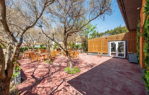 An outdoor seating area with wooden tables and chairs, surrounded by trees and a building with a wooden facade and glass doors.