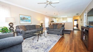 A spacious living room with two grey sofas, a glass coffee table, and a patterned rug. A ceiling fan and a kitchen are visible in the background. - River Oaks Condo, King Suite, Pools & Hot Tub (Myrtle Beach)