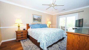 A bedroom with a bed, nightstands, a desk, a ceiling fan, a window, and a painting on the wall. - River Oaks Condo, King Suite, Pools & Hot Tub (Myrtle Beach)