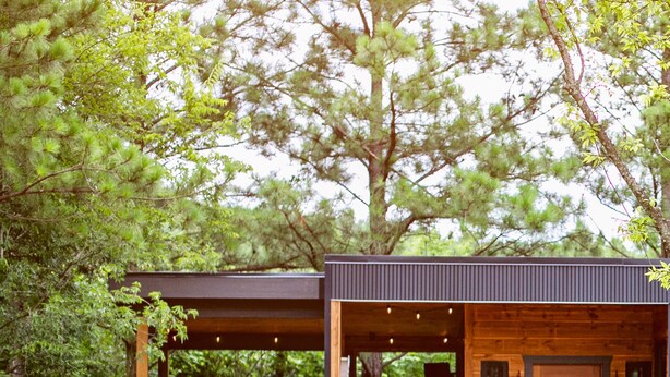 A modern cabin with a covered porch and a deck, surrounded by lush greenery.
