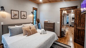A bedroom with a bed, bedside lamps, a dresser, and a view of a kitchen and bathroom through an open door.