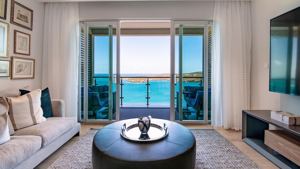 A modern living room with a sofa, coffee table, and a large TV. The room has a sliding glass door leading to a balcony with a view of the sea.