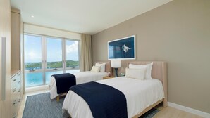 A hotel room with two beds, a large window with a view of the sea and hills, a painting on the wall, and a wooden wardrobe. - Luxe Condo, Ocean Views and pool (St. George)