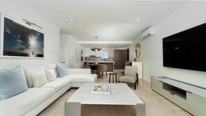 A modern living room with a white sofa, a coffee table, and a TV. There is a kitchen in the background with a dining area. - Luxe Condo, Ocean Views and pool (St. George)