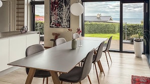 Een moderne eethoek met een grote tafel, stoelen en uitzicht op de zee door grote ramen.