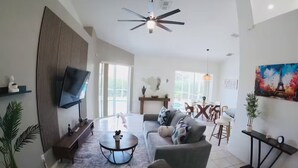 A modern living room with a sofa, coffee table, and TV. There is a ceiling fan, a painting, and a dining area in the background. - Dream Villa W/ Pool Near Disney3 Bed 2 Bath, (Davenport)