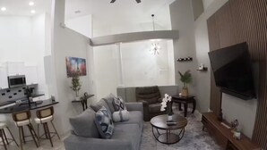 A modern living room with a grey sofa, a coffee table, and a TV. There is a kitchen area with a microwave and a dining table with chairs. - Dream Villa W/ Pool Near Disney3 Bed 2 Bath, (Davenport)