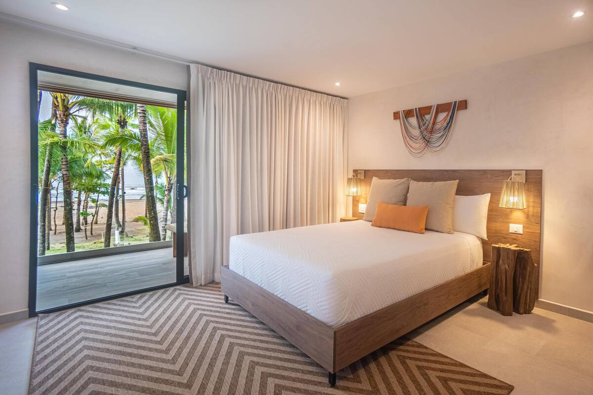 A bedroom with a large bed, a view of palm trees, and a wooden headboard.