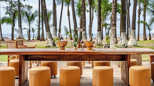 Une terrasse en bois avec une table dressée pour le repas, entourée de tabourets jaunes et moelleux, donnant sur une plage bordée de palmiers.