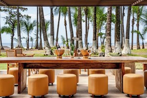 Une terrasse en bois avec une table dressée pour le repas, entourée de tabourets jaunes et moelleux, donnant sur une plage bordée de palmiers.