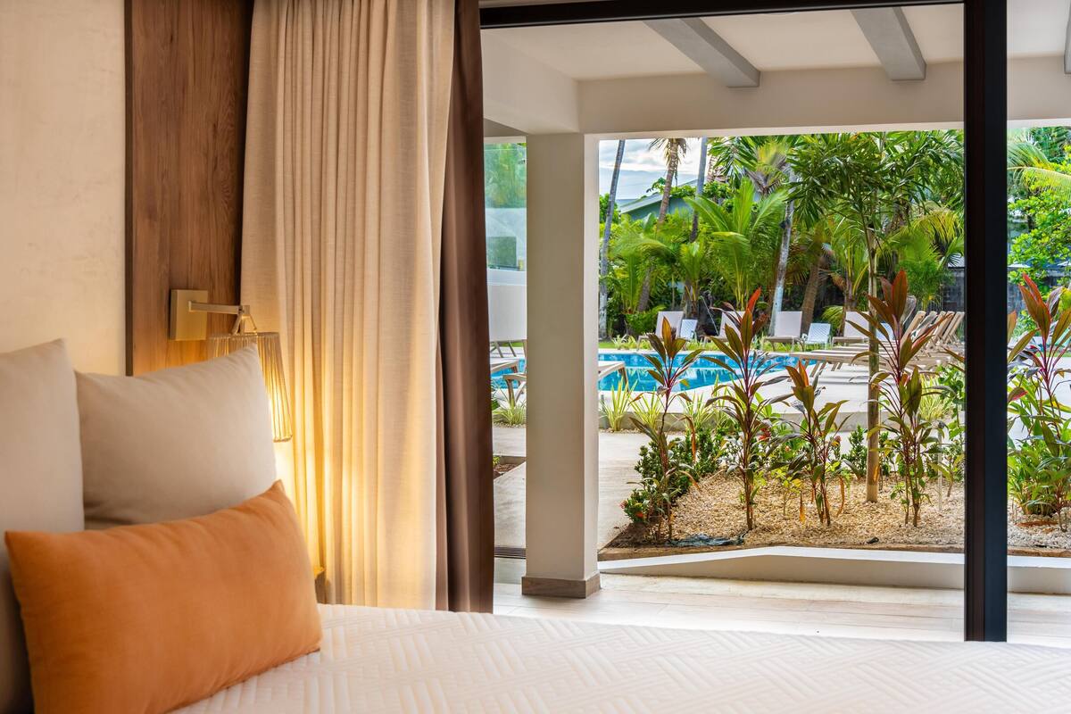 A bedroom with a bed, curtains, and a view of a pool and tropical plants.