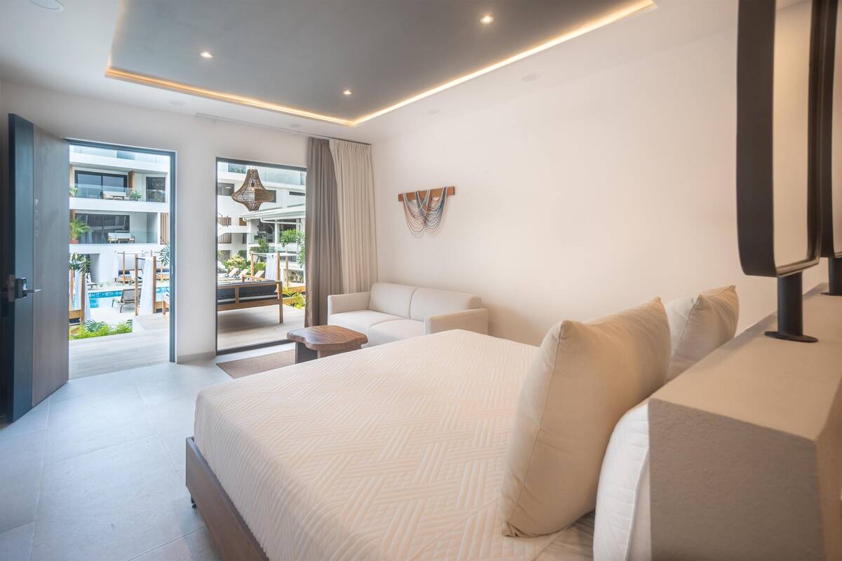 A modern hotel room with a large bed, a sofa, a small table, and a balcony with outdoor furniture.