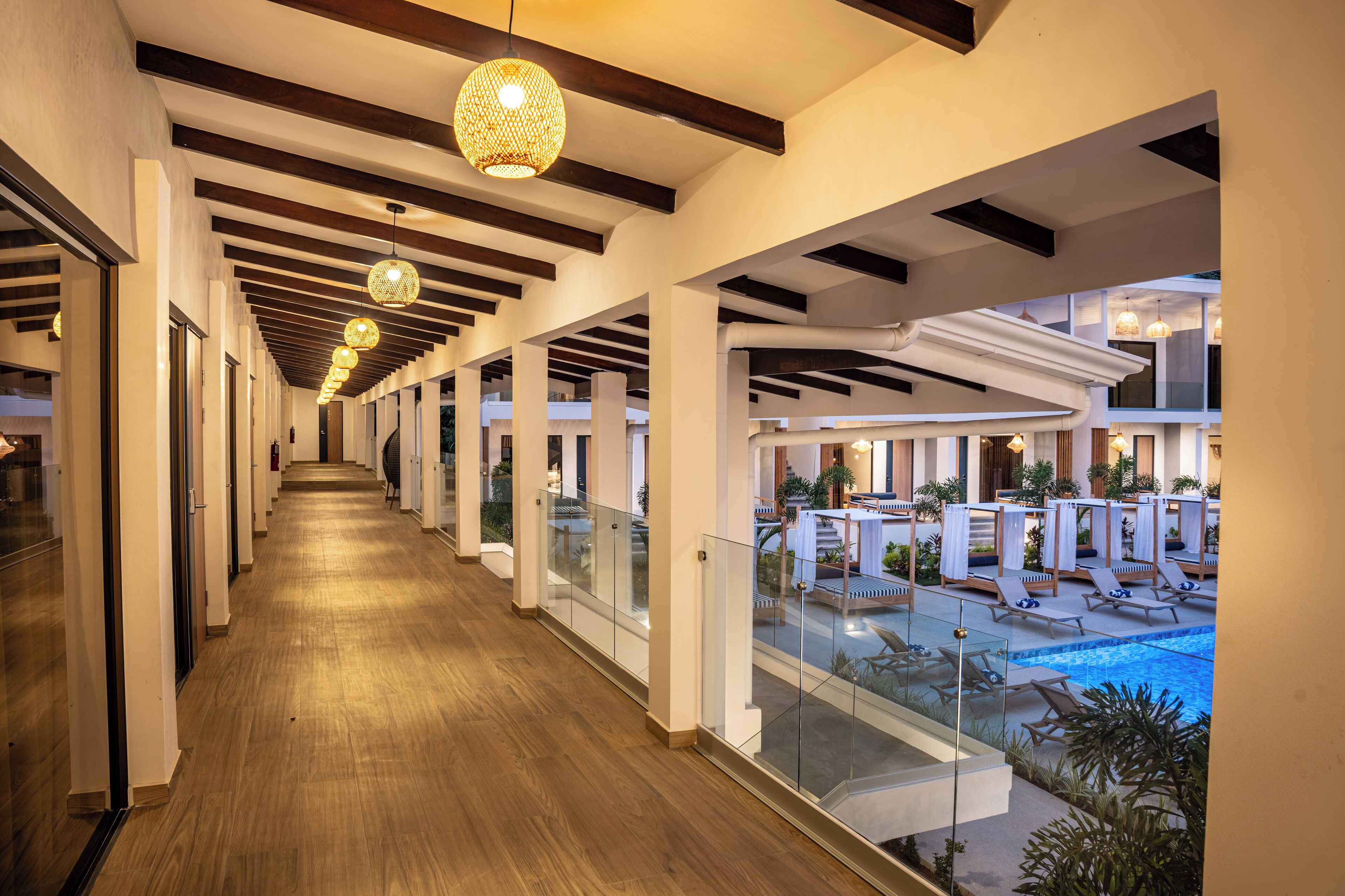 A corridor with wooden flooring and glass railings, leading to a pool area with lounge chairs and umbrellas.