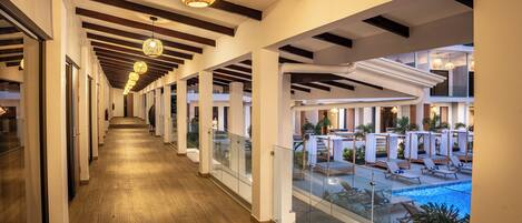 A corridor with wooden flooring and glass railings, leading to a pool area with lounge chairs and umbrellas.