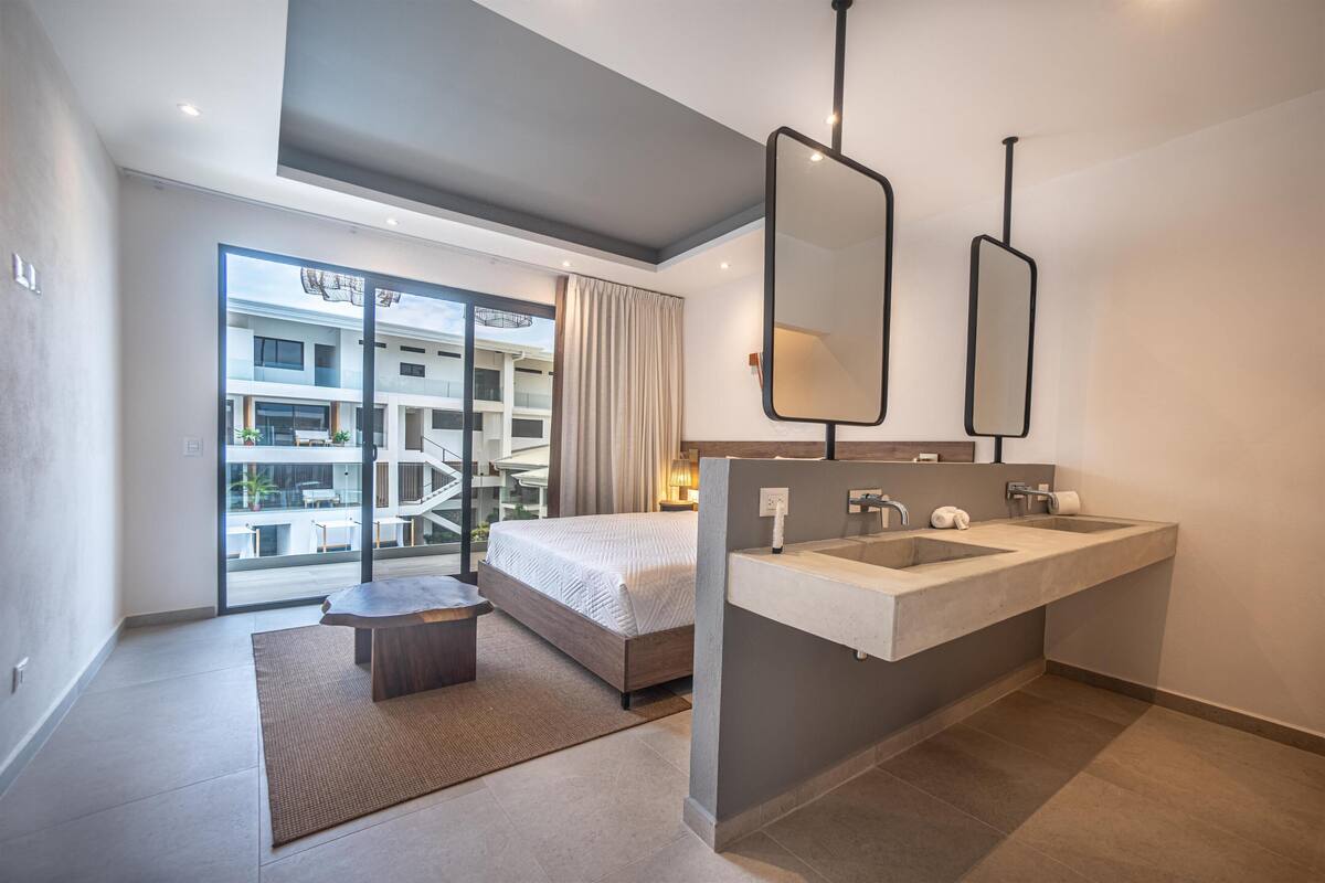A modern hotel room with a large bed, a dual sink vanity, and a view of the building's exterior through a glass door.
