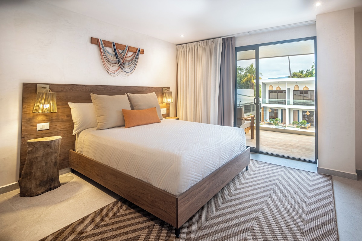 Ein modernes Hotelzimmer mit einem großen Bett, einem Holz-Kopfteil, Nachttischlampen, Blick auf den Balkon und einem großen Fenster.