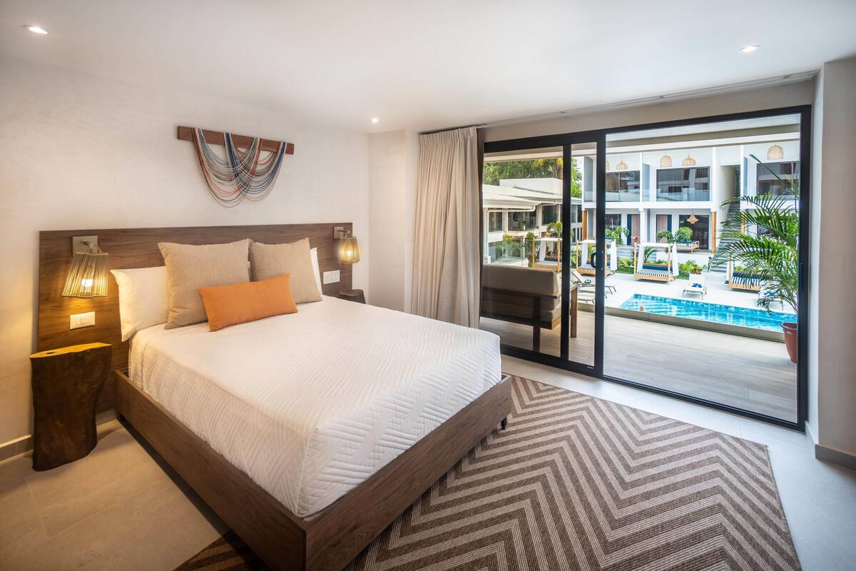 A modern hotel room with a large bed, a view of a swimming pool, and a balcony with outdoor seating.