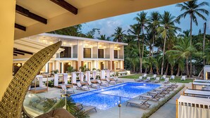 Un espace aménagé au bord de la piscine, avec des chaises longues et une piscine, le tout entouré de palmiers et d’un bâtiment doté de nombreux balcons.