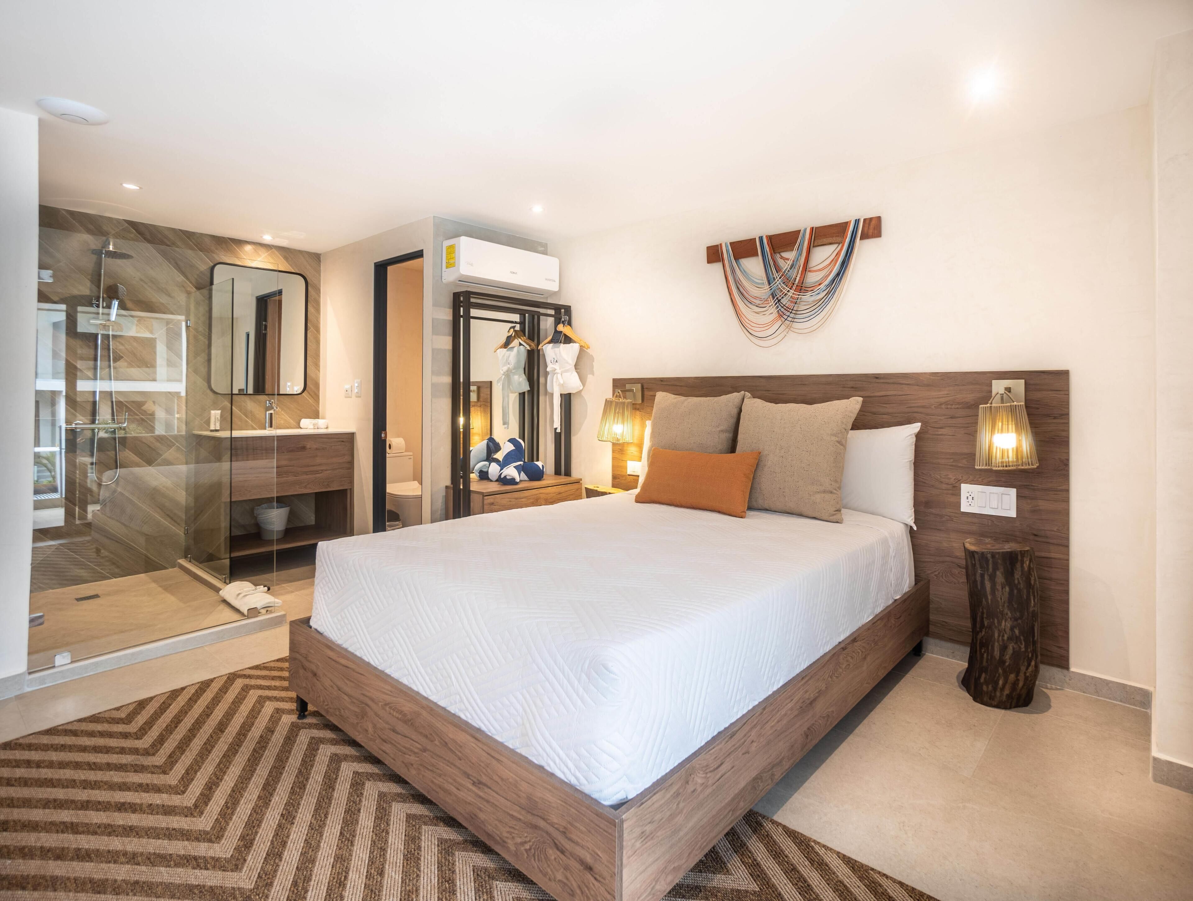 Habitación de hotel moderna con una cama grande, cabina de ducha de vidrio, cabecera de madera y unidad de aire acondicionado montada en la pared.