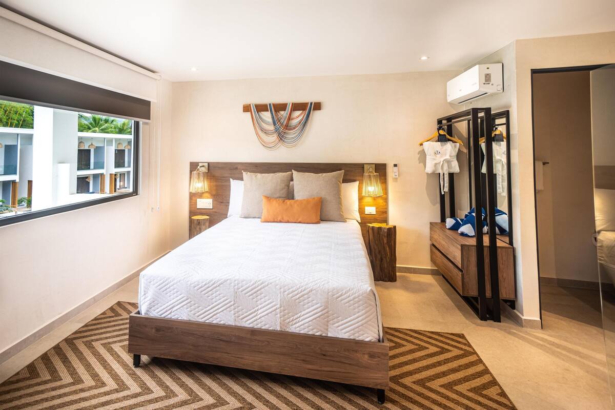A modern bedroom with a large bed, bedside lamps, a wooden headboard, a wardrobe, and a window with a view of greenery.