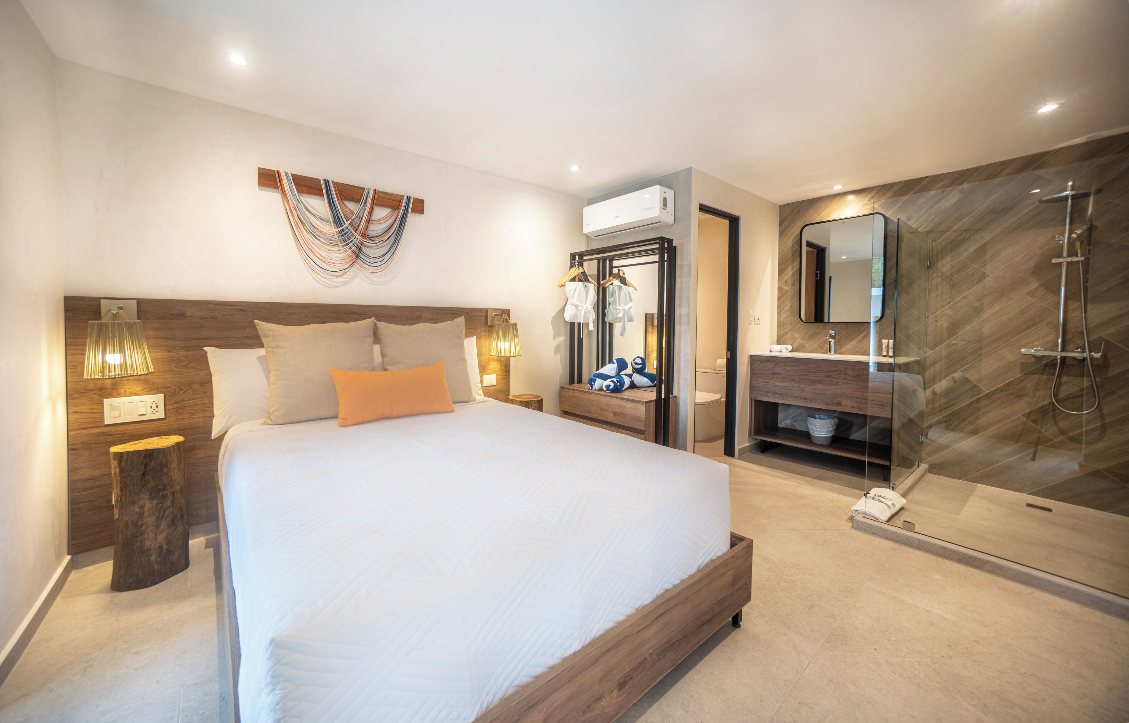 A modern hotel room with a large bed, bedside lamps, a wooden headboard, a mirror, a glass shower enclosure, and a small bedside table.