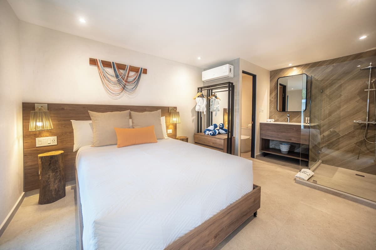 A modern hotel room with a large bed, bedside lamps, a wooden headboard, a mirror, a glass shower enclosure, and a small bedside table.