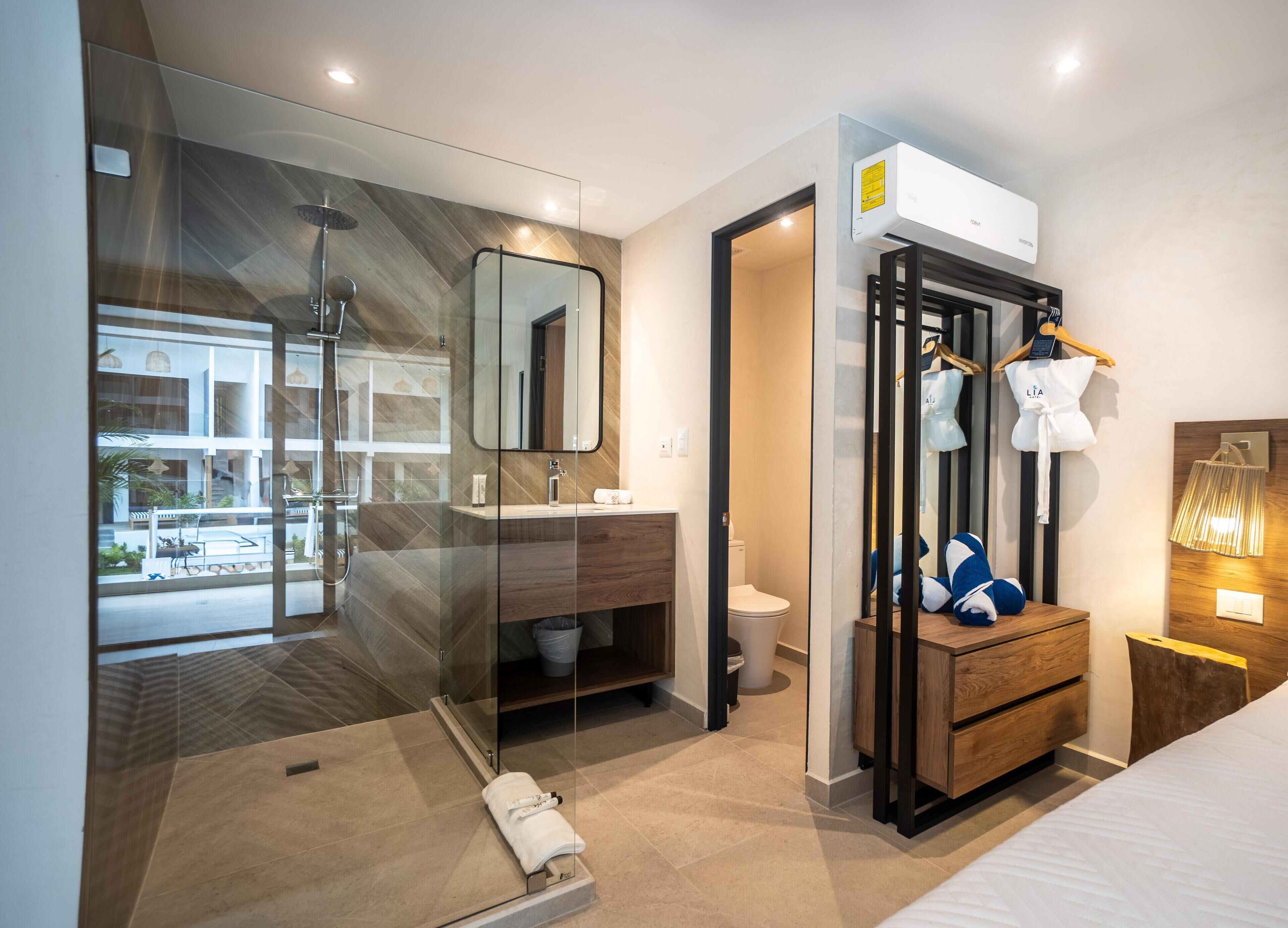 Una habitación de hotel moderna con cabina de ducha de vidrio, tocador de madera, cama y armario con ropa colgada.