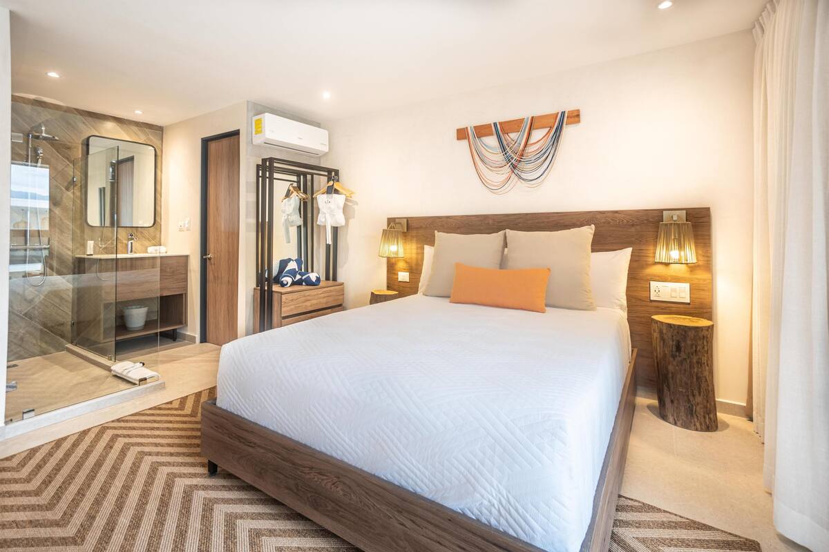 A modern hotel room with a large bed, a glass-enclosed shower, a wooden headboard, and a wall-mounted air conditioner.