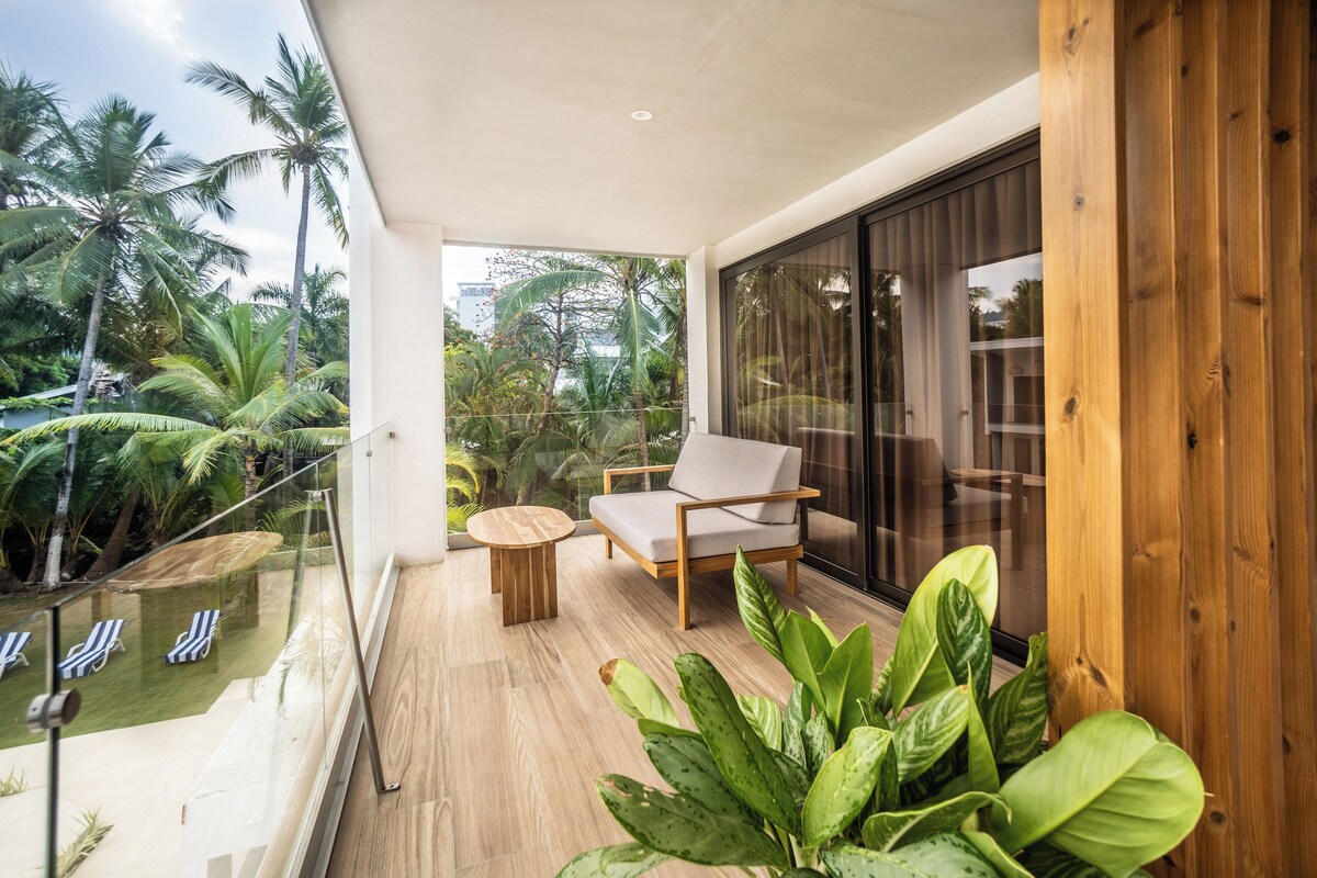 A balcony with a wooden floor, a cushioned chair, a small round table, and a view of palm trees and a pool.