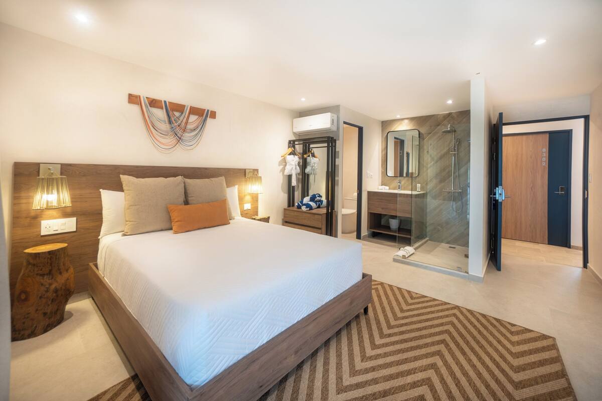 A modern hotel room with a large bed, bedside lamps, a wooden headboard, a glass shower enclosure, and a bathroom with a large mirror and sink.