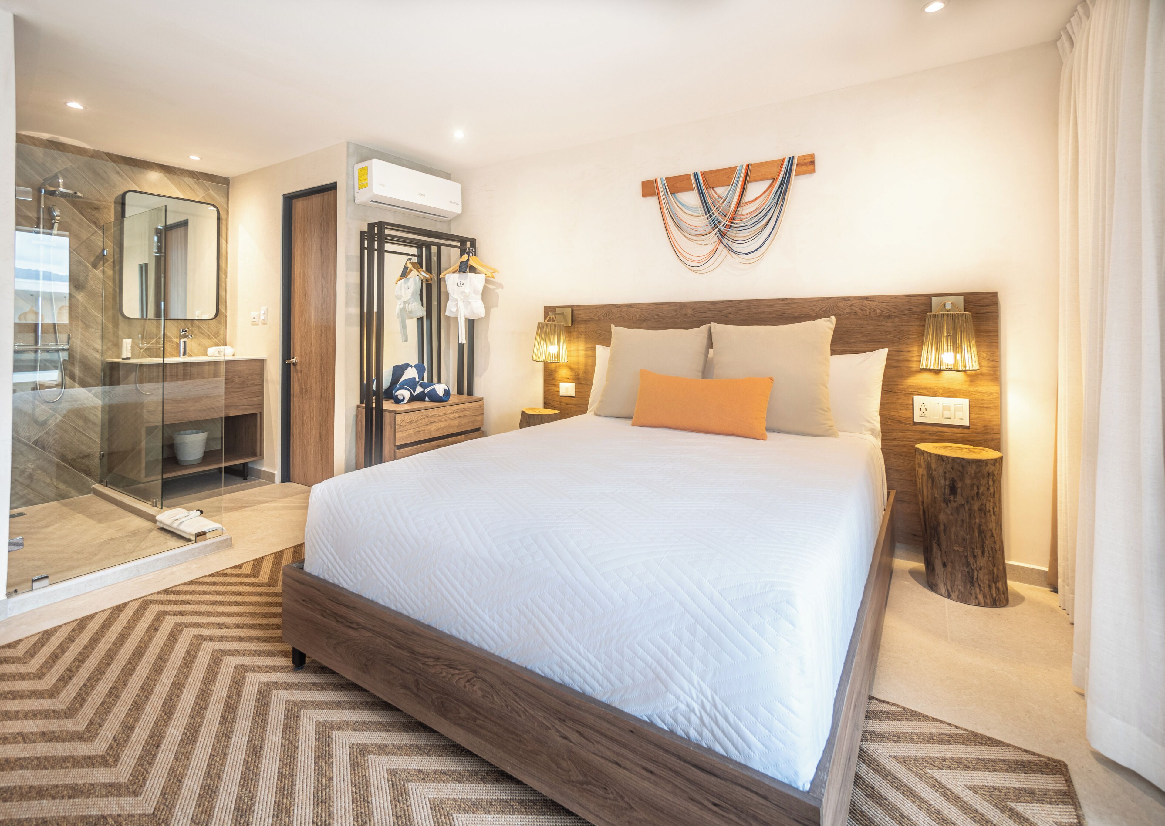 A modern hotel room with a large bed, a glass-enclosed shower, a wooden headboard, and a wall-mounted air conditioner.