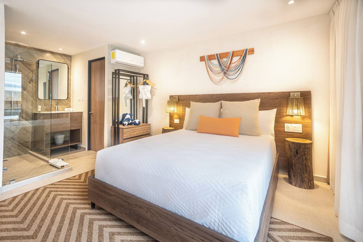A modern hotel room with a large bed, a glass-enclosed shower, a wooden headboard, and a wall-mounted air conditioner.
