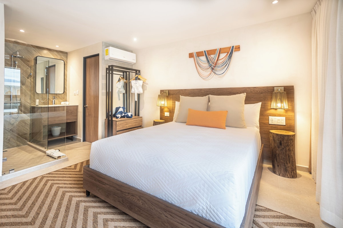 A modern hotel room with a large bed, a glass-enclosed shower, a wooden headboard, and a wall-mounted air conditioner.