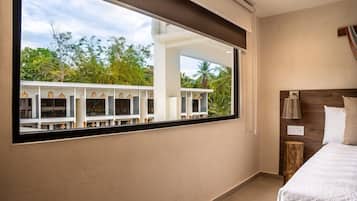 Une chambre avec une grande fenêtre, un lit, une table de chevet et une vue sur un immeuble doté de nombreux balcons et entouré d’arbres.