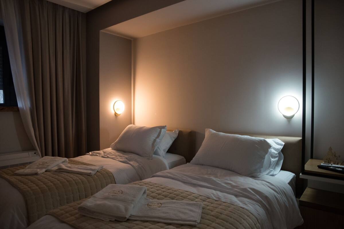 A hotel room with two beds, each with a headboard, and bedside lamps. There are towels on the beds and a nightstand with a remote control.