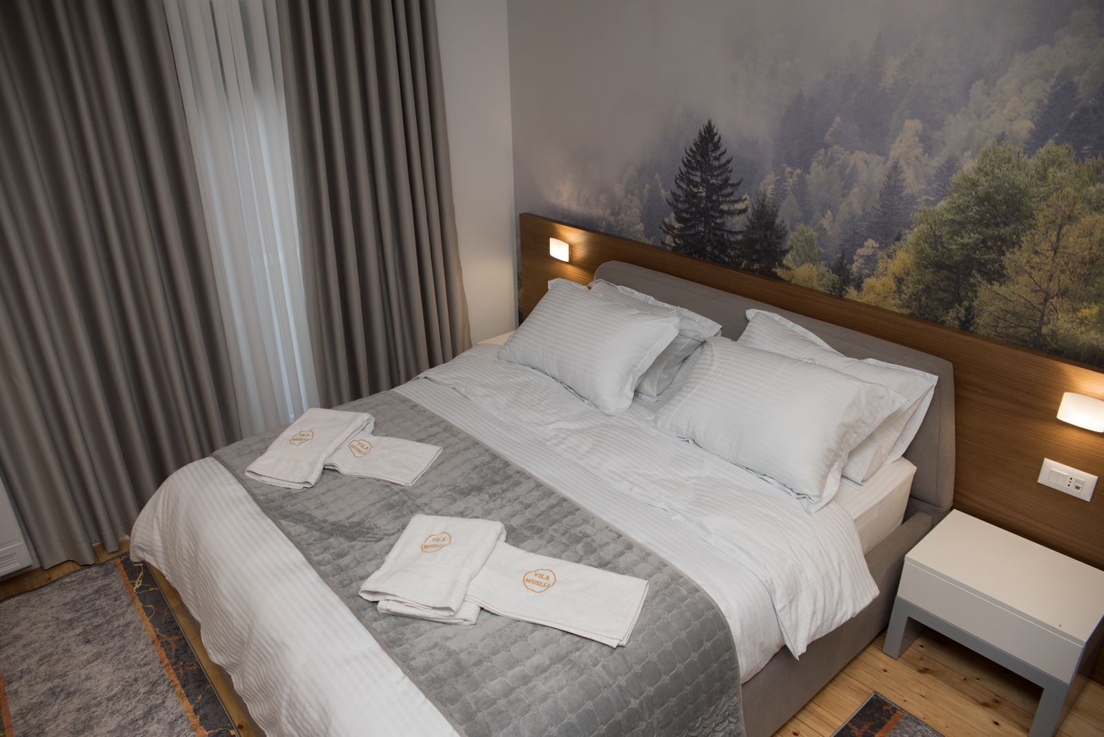 A hotel room with a bed, two bedside tables, a headboard with a forest mural, and a window with curtains.