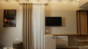 A modern living room with a flat-screen TV, a small white heater, a wooden cabinet, and a large window with vertical blinds. - Vila Muslli (Korçë)