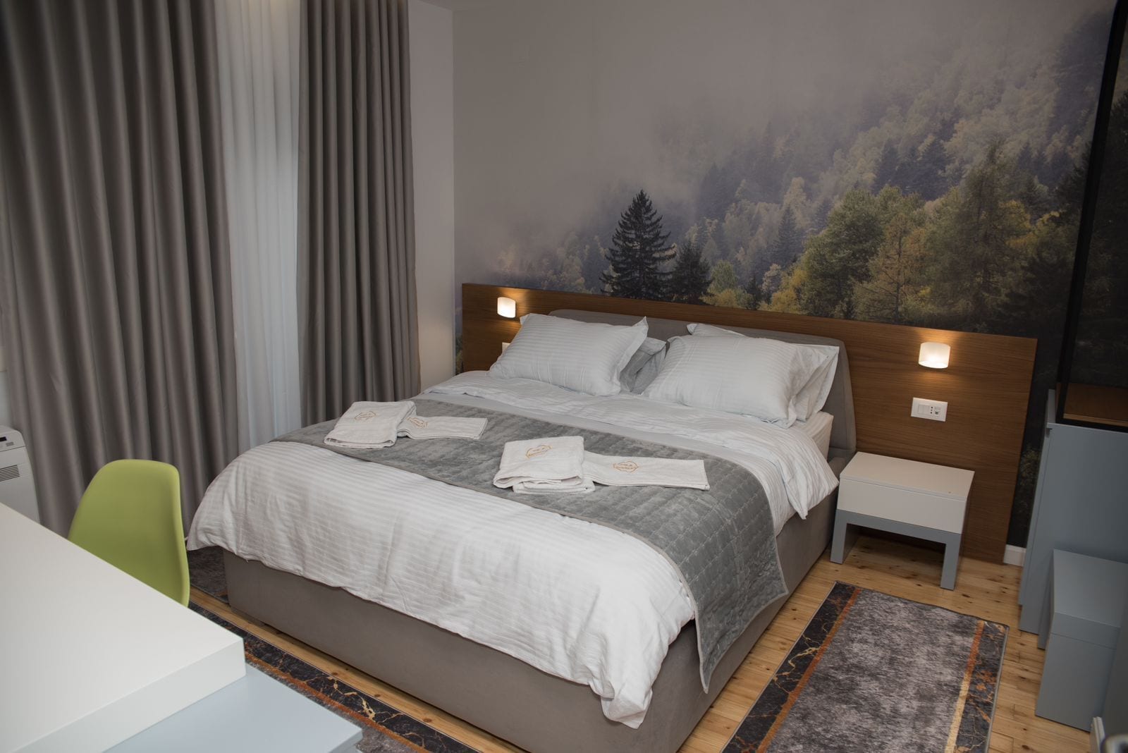 A modern hotel room with a large bed, a desk, and a chair. There is a painting on the wall and a window with curtains.