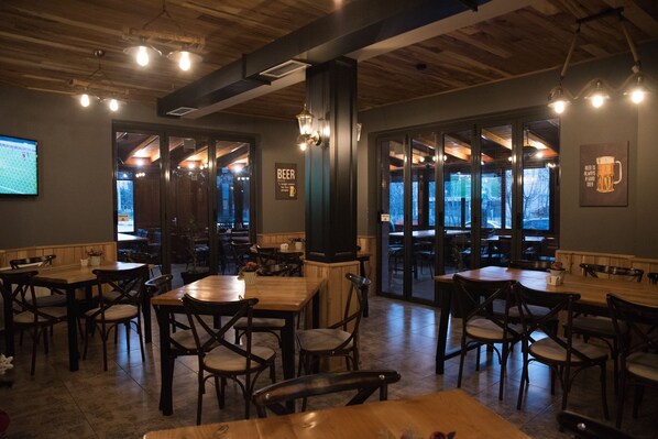 A cozy restaurant with wooden tables and chairs, large windows, and a sign that reads "BEER IS ALWAYS A GOOD IDEA." - Vila Muslli (Korçë)