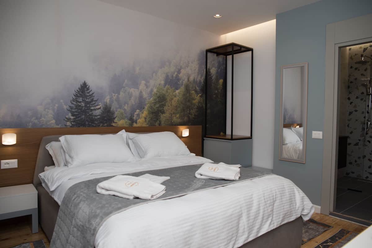 A modern hotel room with a large bed, a headboard with integrated lighting, a large mirror, and a forest-themed wall mural.