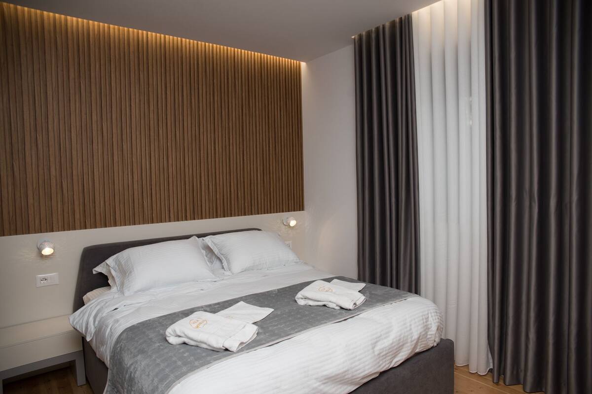 A modern hotel room with a wooden headboard, a large bed with white linens, and a view of a cityscape through sheer curtains.