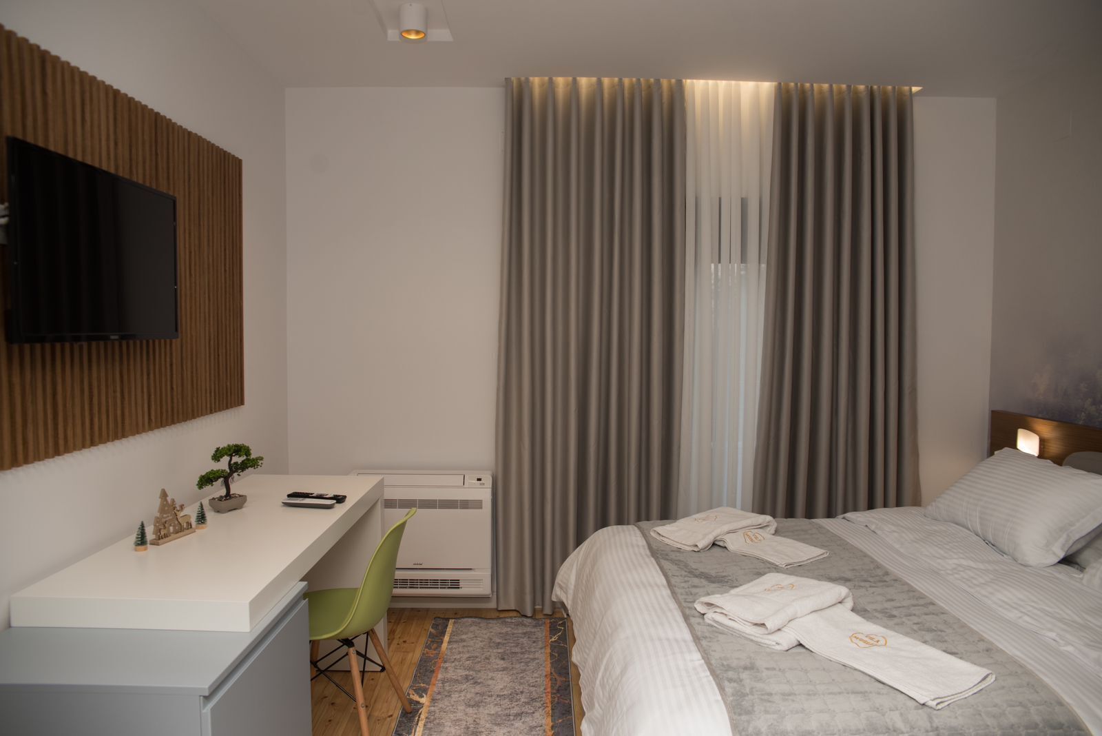 Una habitación de hotel moderna con una televisión de pantalla plana montada en la pared, un escritorio con una silla, una cama con toallas ordenadas y un ventanal con cortinas.