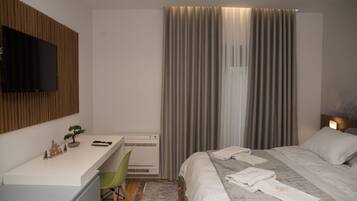 Quarto de hotel moderno com televisão de tela plana na parede, uma escrivaninha com cadeira, cama com toalhas dobradas e uma janela grande com cortinas.
