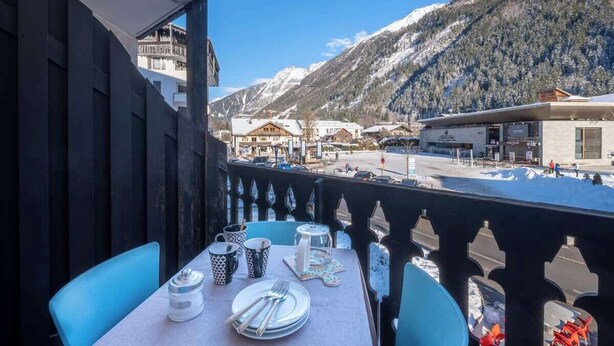 Un balcón con mesa lista para comer, con vistas a un paisaje nevado de montañas y un pueblo en la falda.