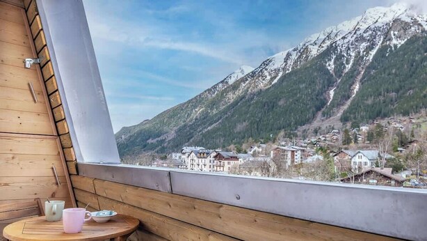 A wooden cabin with a table and chairs, a window with a view of a mountain and a village.
