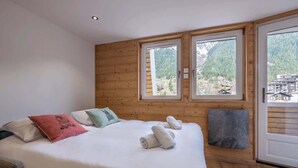 A bedroom with a wooden wall, a bed with white bedding, two windows, and a view of a mountainous area.