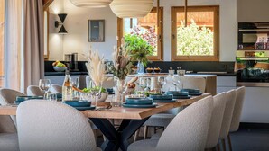A dining area with a wooden table set for six, surrounded by chairs. There are hanging light fixtures above and a window with a view of greenery.