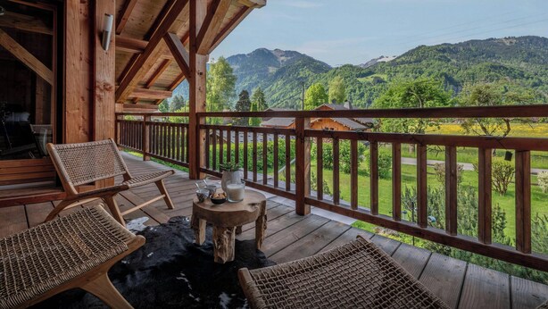 A wooden deck with wicker furniture, a small table with a cup and saucer, and a view of green hills and a house in the distance.