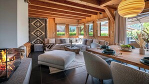 A cozy living room with a fireplace, wooden beams, a sofa set, and a dining area with a view of greenery outside.